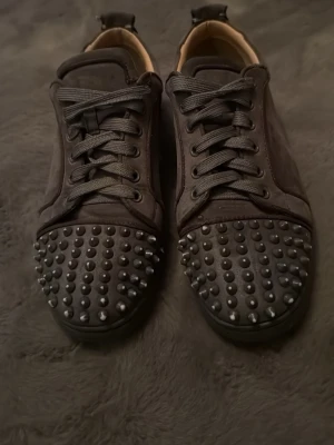 Svarta sneakers med nitar från Louboutin - Unika svarta sneakers från Christian Louboutin med gråa nitar på tån och klassisk röd sula. Skorna har snörning och är tillverkade i mocka med läderdetaljer. Perfekta för dig som vill sticka ut med en edgy och exklusiv stil.