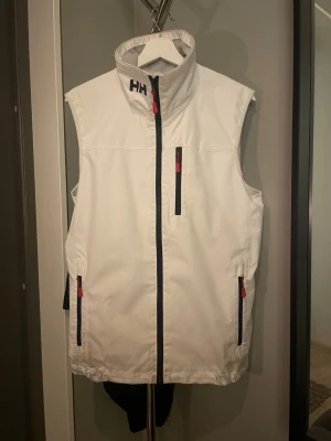 Vit Helly Hansen väst med dragkedjor - Snygg vit väst från Helly Hansen med hög krage och svarta detaljer. Västen har tre dragkedjeförsedda fickor med röda dragare och HH-logga på kragen och ryggen. Perfekt för lager-på-lager och outdoor-stil. Jag har kvittot och de grejer som följde med till den. Bara att höra av sig vid frågor!