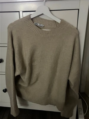 Beige stickad tröja från Zara - Mysig beige stickad tröja från Zara med rund halsringning och ribbade muddar vid ärmslut och nederkant. Tröjan har en avslappnad passform och är perfekt för lager-på-lager-stil under kyliga dagar. storlek S.
