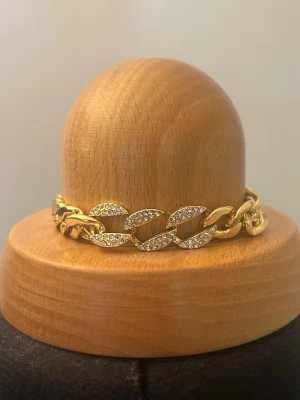 Guldigt kedjearmband med strass - Snyggt armband i guldfärgad metall med breda länkar, där vissa länkar är dekorerade med glittrande strass. Justerbar längd med förlängningskedja och liten kula i slutet. Perfekt för dig som gillar statement-smycken och vill ha något som syns.