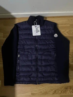Mörkblå dunväst från Moncler - Snygg mörkblå dunväst från Moncler med stickade ärmar och hög krage. Västen har två dragkedjefickor fram och en Moncler-logga på vänster ärm. Framsidan är quiltad och baksidan är helt stickad för en modern look.