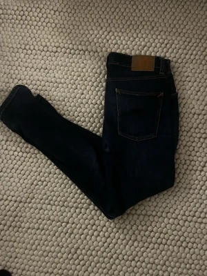 Mörkblå jeans från Nudie Jeans - Snygga mörkblå jeans från Nudie Jeans med klassisk femficksmodell och kontrastsömmar. Jeansen har en rak passform och är tillverkade i slitstark bomullsdenim. Perfekta för dig som gillar stilrena och tidlösa jeans.