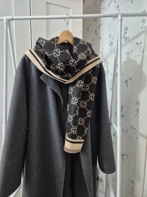 Svart halsduk med beige mönster - Svart halsduk med beige blommönster och ränder längs kanterna. Halsduken är rektangulär och stickad i ett mjukt material som känns skönt runt halsen. Perfekt för dig som vill ha en trendig accessoar med grafisk design.