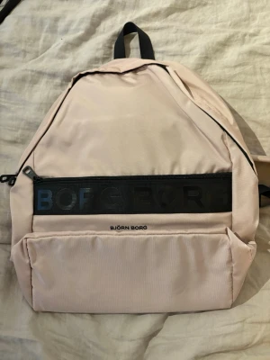 Ljusrosa Björn Borg ryggsäck - Snygg ljusrosa ryggsäck från Björn Borg med svart dragkedja och logga framtill. Väskan har ett stort huvudfack, en framficka med dragkedja och justerbara axelband i svart mesh. Perfekt för skola eller cityhäng.