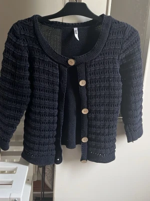 Mörkblå stickad kofta med träknappar - Snygg mörkblå stickad kofta med strukturmönster och fyra träfärgade knappar framtill. Koftan har rund halsringning och trekvartsärmar. Perfekt att slänga över en topp för en chill och stilren look.