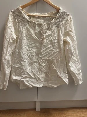 Vit odd molly med knyt och smock - Säljer en vit blus från Odd Molly med smockdetaljer vid axlarna och knytband med tofs framtill. Blusen har lång ärm och är gjord i mjuk bomull, perfekt för en avslappnad och trendig look.