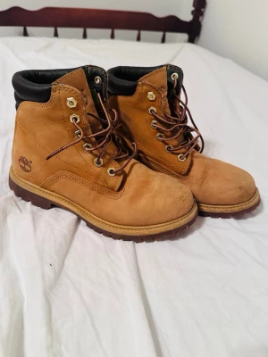 Timberland klassiska kängor mocka brun - Säljer ett par klassiska Timberland kängor i brun mocka med svart vadderad kant och robust sula. Kängorna har snörning med metallhål och ikonisk Timberland-logga på sidan. Perfekta för dig som vill ha en tidlös och snygg look.