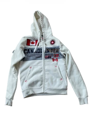 Vit hoodie från Napapijri med tryck - Snygg vit hoodie från Napapijri med dragkedja, huva och flera tryckta flaggor samt logga på bröstet. Stora fickor framtill och detaljer i rött. Perfekt för dig som gillar streetwear och vill sticka ut med coola detaljer.