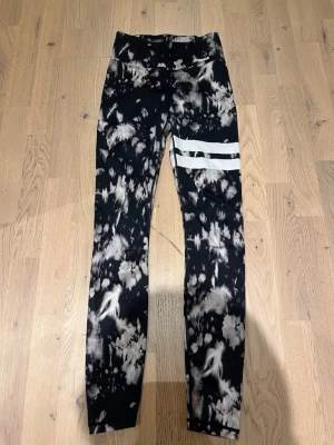 Svarta tie-dye leggings från Stronger - Snygga svarta leggings från Stronger med tie-dye mönster i vitt och två vita ränder på ena benet. Hög midja och stretchigt material som passar perfekt till träning eller chill. Logga på höften och bekväm passform.