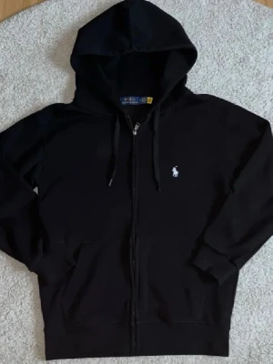 Svart hoodie från Polo Ralph Lauren - Klassisk svart hoodie med dragkedja från Polo Ralph Lauren. Har huva med dragsko, två fickor framtill och den ikoniska vita logon broderad på bröstet. Perfekt för en avslappnad och stilren look. Tillverkad i mjukt bomullsmaterial.