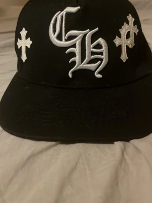 Svart keps med vita kors och brodyr - Säljer en svart keps med platt skärm och flera vita kors broderade runtom. Framtill finns stora broderade bokstäver i vitt. Kepsen har en modern streetwear-look och är tillverkad i ett klassiskt bomullsmaterial.