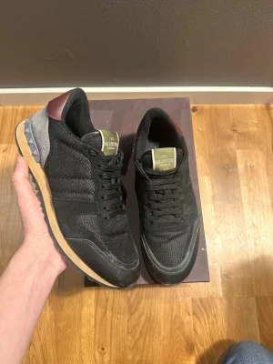 Valentino Garavani svarta sneakers - Svarta Valentino Garavani sneakers med mesh och mockadetaljer, olivgröna tungor med logga och vinröda hälpartier. Snygg platt sula med nitar på bakkappan och klassisk snörning. Perfekta för dig som vill ha en exklusiv och stilren look.