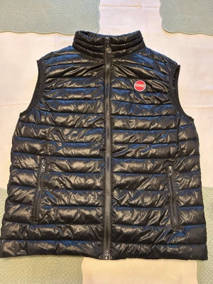 Svart dunväst från Colmar - Snygg svart dunväst från Colmar med quiltad design och hög krage. Västen har två dragkedjeförsedda fickor och en diskret Colmar-logga på bröstet. Perfekt att slänga över en hoodie eller tröja för extra värme. Materialet är glansigt och lätt, vilket ger en modern look. Storlek på taggen är xxl men den passar L