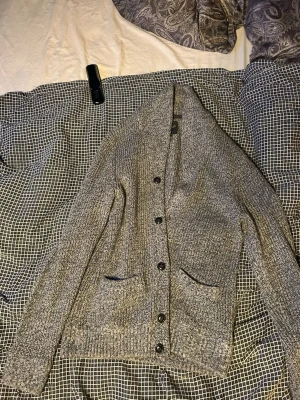 Grå stickad kofta från Mario Conti - Mysig gråmelerad stickad kofta från Mario Conti med sjalkrage och knäppning framtill. Två fickor framtill och ribbade muddar vid ärmslut och nederkant. Perfekt för lager på lager och chill stil.