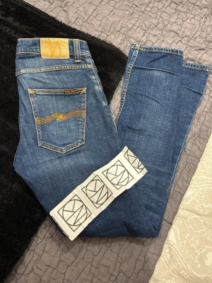 Blå jeans från Nudie Jeans - Snygga blå jeans från Nudie Jeans med klassisk femficksdesign och gulddetaljer på bakfickan. Jeansen har normal passform och raka ben. Tillverkade i slitstarkt bomullsjeansmaterial, perfekta för en avslappnad stil. Grim Tim slim fit