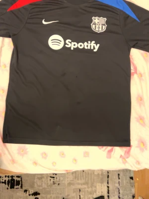 FC Barcelona svart matchtröja Nike - Svart FC Barcelona matchtröja från Nike med klubbmärke och Spotify-logga på bröstet. Tröjan har röda och blå detaljer på axlarna och är tillverkad i ett lätt, sportigt material som passar perfekt till fotbollsträning eller match.