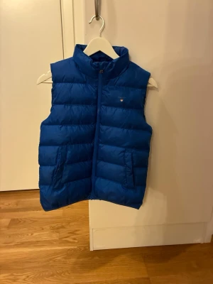Blå dunväst från Gant -  Snygg blå dunväst från GANT med quiltad design och hög krage. Västen har två praktiska framfickor och stängs med dragkedja framtill. Den diskreta GANT-loggan på bröstet ger ett stilrent och modernt uttryck. Perfekt att bära över en hoodie eller tröja för extra värme utan att begränsa rörelsefriheten.  Unisexmodell i storlek 146/152, tillverkad i polyester. En mångsidig väst som passar lika bra till vardag som till olika aktiviteter under flera årstider.
