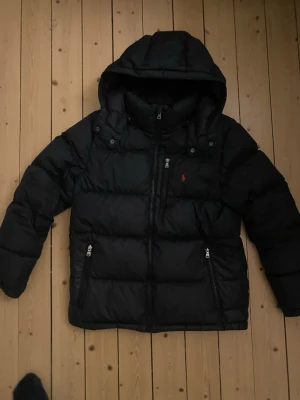 Svart dunjacka från Polo Ralph Lauren - Storleken är 14-16år Svart puffer dunjacka från Polo Ralph Lauren med huva och dragkedja framtill. Jackan har två fickor med dragkedja och den klassiska röda Polo-loggan på bröstet. Perfekt för kalla dagar och riktigt snygg streetstil.