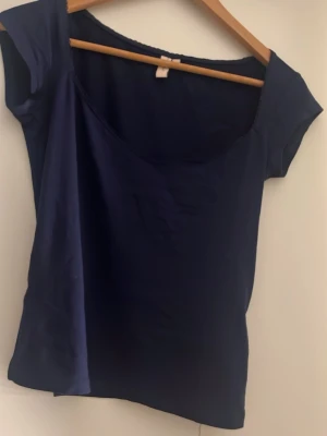 Mörkblå topp från Nelly - En enkel och stilren mörkblå topp från Nelly med korta ärmar och rundad, djup urringning. Tillverkad i mjuk bomullsmix som känns skön mot huden. Perfekt att matcha med jeans eller kjol för en clean look. Aldrig använd