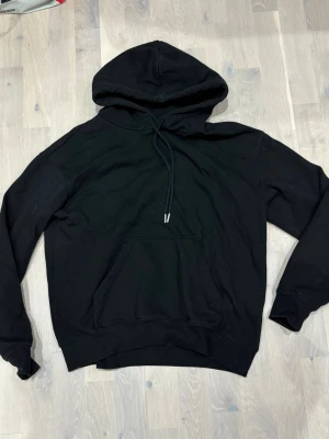 Svart hoodie med huva och magficka - Klassisk svart hoodie med stor huva och dragsnören. Hoodien har en rymlig magficka framtill och ribbade muddar vid ärmslut och nederkant. Tillverkad i mjukt material, perfekt för en avslappnad stil.