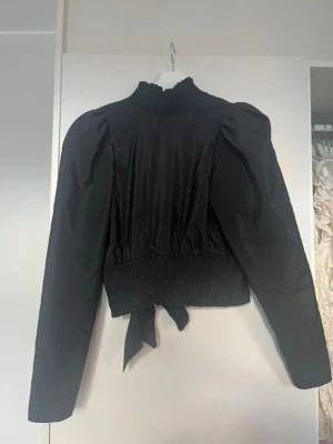 Svart blus med puffärmar och hög krage - Svart blus med puffiga långärmar, öppen rygg, smockad hög krage och bred smock i midjan. Materialet har en lätt glansig finish och baktill finns en knytning i midjan med den öppna ryggen ⭐️