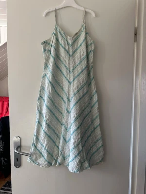 Randig sommarklänning med smala axelband - Luftig långklänning med smala axelband i vitt och ljusblått randigt mönster. Klänningen har v-ringning, dragkedja i ryggen och är tillverkad i ett mjukt bomullsliknande material. Perfekt för varma dagar och har en avslappnad passform.