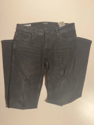 Svarta ripped jeans Jack & Jones - Säljer ett par svarta jeans från Jack & Jones med slitna detaljer och hål på benen. Jeansen har fem fickor, normal passform och klassisk knappgylf. Perfekta för dig som gillar en edgy stil och vill ha något som sticker ut.