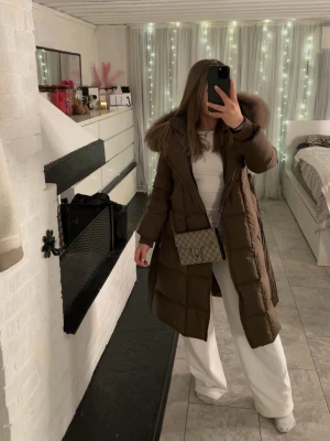 Brun lång dunjacka med pälskrage - Snygg lång brun dunjacka med stor pälskantad huva och quiltad design. Jackan har dragkedja framtill och en rak passform som ger en chill och modern look. Perfekt för kalla dagar när du vill vara både varm och stilren. Köpt från Feminina of Sweden, Casablanca, använt ca 2-3 månader köpte för 4 tusen 