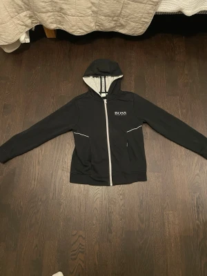 Svart hoodie från Hugo Boss - Svart hoodie från Hugo Boss med vit dragkedja och logga på bröstet. Utrustad med huva och vita detaljer vid fickorna. Insidan av huvan är ljusgrå. Perfekt för en avslappnad och stilren look.