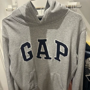 Grå hoodie från GAP med stor logga - Säljer en klassisk grå hoodie från GAP med stor, mörkblå broderad logga på bröstet. Tröjan har huva och en stor magficka framtill. Perfekt för chill dagar och enkel att matcha med jeans eller joggers. Basic hoodie! Fråga gärna om fler bilder! Kom gärna med förslag på pris !