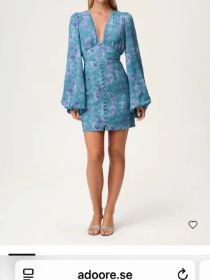Blommig blå kortklänning från Adoore - Supersnygg blå kortklänning från Adoore med lila blommönster, v-ringning och långa puffärmar. Klänningen har knappar framtill och markerad midja som ger en feminin siluett. Perfekt för vår och sommar!ANVÄND 0 GGR MED PRISLAPP KVAR!!!!!pris kan diskuteras kom med förslag 