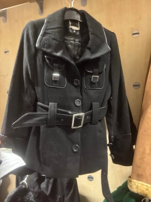 Svart kavajjacka med bälte - Alla storlekar finns. Pris kan diskuteras. Perfekt inför våren. Stockholms stil.                                 Stilren svart kavajjacka från Saint Clair med markerad krage, stora knappar och dekorativa fickor framtill. Jackan har ett brett bälte i midjan med metallspänne och quiltat innerfoder. Långa ärmar och detaljer i silver ger en cool touch.