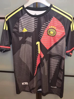 Neuer Adidas - Snygg svart målvaktströja från Adidas med Tysklands landslagsmärke och fyra stjärnor på bröstet. Tröjan har gula och röda detaljer, samt texten 'NEUER' och nummer 1 på ryggen. Tillverkad i funktionsmaterial med HEAT.RDY-teknologi. Den är tvättad men oanvänd. Tveka inte för att ställa frågor! Priset är inte hugget i sten.