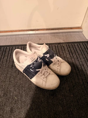 Vita Valentino sneakers med blå detaljer - Valentinos väldigt bra skick en tvätt o putsa så är den som nya, knappt använda ca 5 gånger då dem är lite små därav slitning i häl. Säljer ett par vita Valentino sneakers med bred svart rem över vristen och nitar som ger en cool edge. Skorna har rund tå, platt sula och klassisk snörning. Perfekta för dig som vill ha något stilrent men ändå sticker ut.