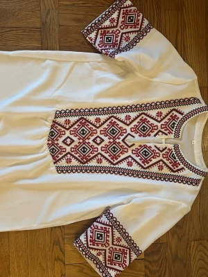 Vit skjorta med röd brodyr - Unik vit skjorta med detaljerad röd och svart broderad design på bröstet och ärmarna. Skjortan har halvlång ärm och knappar vid halsen. Perfekt för dig som gillar traditionella mönster med modern twist.