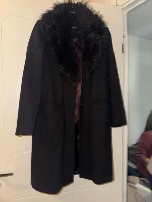 Svart lång kappa  - Svart kappa med fusk päls framtill, två fickor och en knapp för att stänga kappan om man så önskar.