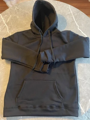 Svart hoodie med magficka och huva - Stilren svart hoodie med magficka och huva helt oanvänd i medium fit tillverkad av 100% polyester för hög komfort och hållbarhet. Mjuk, lätt och behåller passformen tvätt efter tvätt. Perfekt för både vardag och fritid.🌟