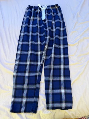 Blå-rutiga pyjamasbyxor med rosett - Mysiga pyjamasbyxor i blått rutigt mönster med vita och svarta detaljer. Byxorna har en bred resår i midjan och en söt vit rosett framtill. Perfekta för chillkvällar hemma.