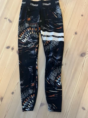 Mönstrade tights från Stronger - Snygga svarta tights från Stronger med färgglatt abstrakt mönster i vitt, orange och grått. Hög midja med logotypen 'STRONGER' tryckt runt linningen. Tillverkade i stretchigt funktionsmaterial som passar perfekt till träning.