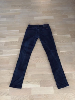Mörkblå jeans från Nudie Jeans - Säljer ett par mörkblå jeans från Nudie Jeans med klassisk femficksmodell och raka ben. Jeansen har snygga sömdetaljer på bakfickorna och en svart patch i midjan. Perfekta för dig som gillar stilrena och tidlösa jeans.