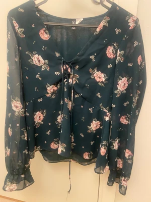 Blommig blus med snörning från H&M - Supersöt blommig blus från H&M i mörkgrönt med rosa och vita rosor. Blusen har v-ringning med snörning framtill, långa transparenta ärmar med resår vid ärmslut och ett flowy, lätt material som ger en chill vibe. Perfekt för dig som gillar romantiska detaljer och mjuka färger.