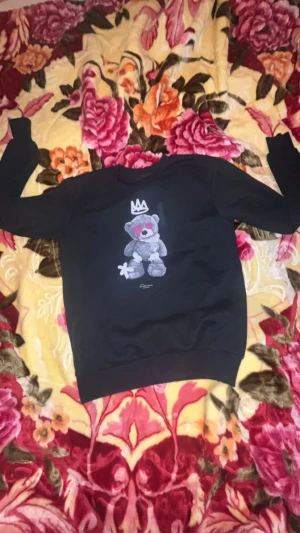 Svart sweatshirt Closure London med nalle - Svart sweatshirt från Closure London i storlek S med en stor tryckt nalle på framsidan, detaljerad med krona och rosa markeringar. Tröjan har rund hals och långa ärmar, tillverkad i mjuk bomullsmix som är skön att bära. Perfekt för dig som gillar streetwear och coola prints.