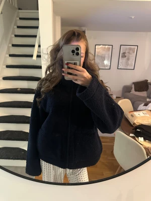 Mörkblå teddyjacka med dragkedja - Säljer en mörkblå teddyjacka med oversized passform och hög krage. Jackan har en grov dragkedja framtill och två stora fickor. Materialet är fluffigt fleece som känns riktigt mjukt och cozy. Perfekt för kalla dagar när man vill vara både chill och snygg.