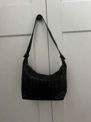 Svart flätad axelväska i skinn - Snygg svart axelväska med flätad design och justerbart axelband. Väskan är tillverkad i skinn och har en silverfärgad dragkedja upptill. Perfekt storlek för att få plats med allt du behöver och stilren look som passar till många outfits.