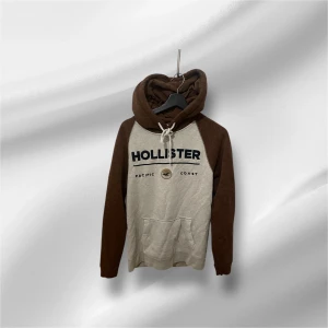 Beige och brun hoodie från Hollister - Snygg hoodie från Hollister i beige med bruna ärmar och huva. Stor logotyp framtill, snörning vid halsen och stor magficka. 