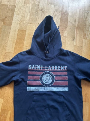 Mörkblå Saint Laurent hoodie. Pris kan diskuteras  - Snygg mörkblå hoodie från Saint Laurent med tryck i rött, vitt och blått framtill. (Vet ej om de är äkta köpt på Vinted för 1500 tröjan är också liten i storlek passar xs och S)