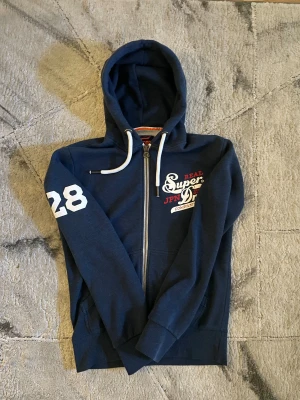 Superdry Zip Hoodie - Oversized XS hoodie i blå. Jätte skön och varm hoodie. Så gott som ny! Inga fläckar,hål,urtvättningar eller missfärgningar. Den är XS men hade passat S eftersom att modellen är lite oversized (passar mig som bär XS men bara lite stor). EJ tillgänglig i affärer längre. Axel brett 49 cm & längd från axeln ner 66 cm. Ny tvättad! OBSERVERA alla bilder. Priset kan diskuteras vid snabbt köp och skickas inom 24h