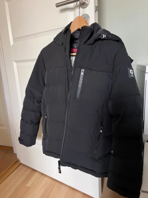 Svart pufferjacka från Garcia stl 152/158 - Svart pufferjacka från Garcia med huva och flera dragkedjeförsedda fickor. Jackan har quiltad design, lång ärm och reflekterande detalj på huvan. Passar ca 12år. Knappt använd. 