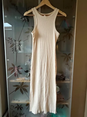 Ribbad långklänning från H&M - Säljer en ribbad, ärmlös långklänning från H&M i ljusbeige. Klänningen har rund halsringning, slits på sidan och är tillverkad i mjuk bomullsmix. Perfekt för en clean och stilren look.