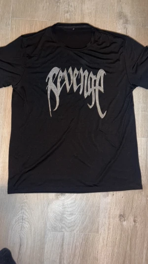 Svart Revenge t-shirt med tryck - Svart t-shirt från Revenge med stort silvrigt tryck på bröstet i coolt, grafiskt typsnitt. Klassisk rund hals och korta ärmar. Mjuk och lätt bomullskvalitet som känns skön mot huden. Perfekt för dig som gillar streetwear och vill sticka ut.